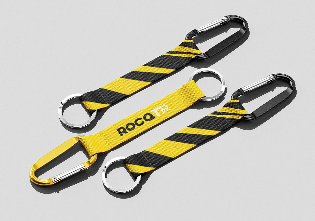Porte-clés mousqueton en sangle tissée jaune et noir – objet promotionnel avec logotype ROCQTR, motif de bandes diagonales et jaune plein, déclinaison d'identité visuelle sur produit dérivé